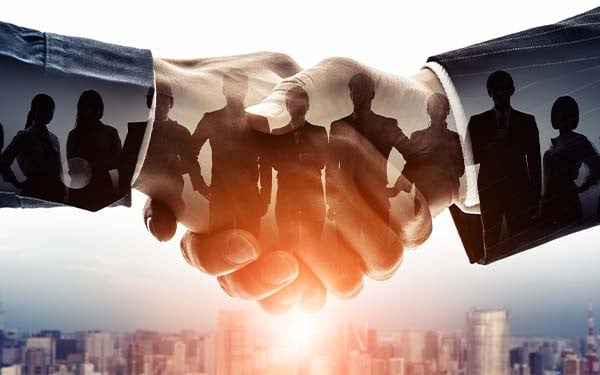 Digital composite image of two hands shaking, containing the shadow of businesspeople above a city skyline.